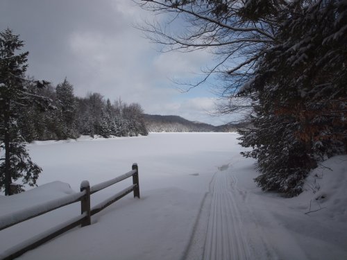 A snowy landscape with a frozen lake, surrounded by trees and mountains under a partly cloudy sky.