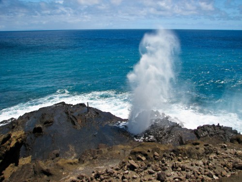 A water spout erupts from rocky shore, with waves crashing against the coastline and a blue ocean in the background.