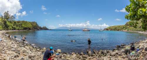 A panoramic view of a calm bay with people swimming, rocky shores, and boats in the distance under a clear blue sky.