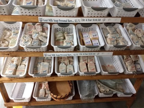 Shelves displaying various goat milk soap personal bars in baskets, with pricing signs for individual and bulk purchases.