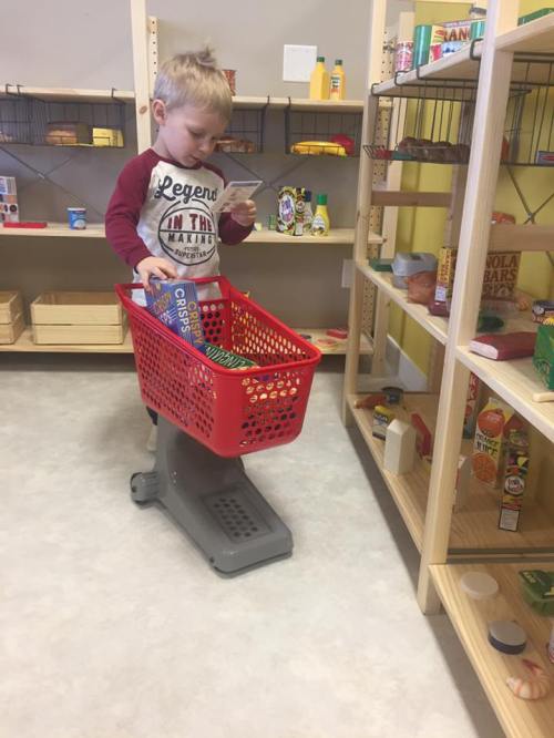 A young child stands with a small shopping cart, holding a snack box in a play grocery store setting.