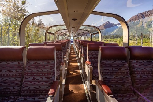 Interior of a scenic train with empty rows of seats and large windows showcasing a mountainous landscape outside.