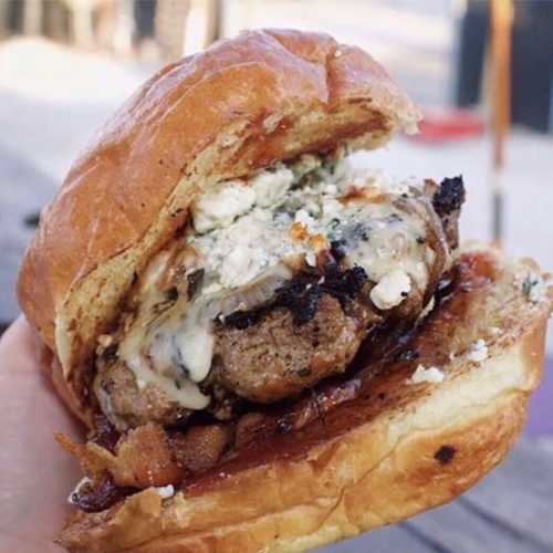 A close-up of a juicy burger with melted cheese, crumbled blue cheese, and toppings, held in a hand.