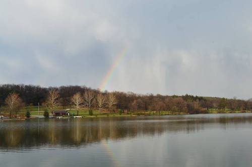 A serene lake reflects a rainbow and trees, with a cloudy sky in the background.