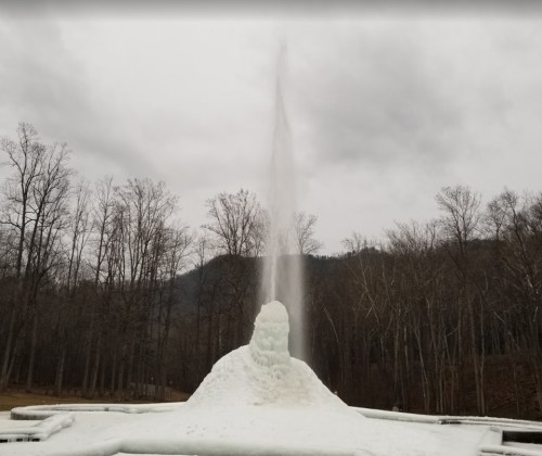 A frozen fountain with a tall spray of water, surrounded by bare trees and mountains under a cloudy sky.
