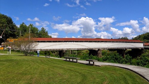 A white covered bridge spans a river, surrounded by green grass and trees under a blue sky with fluffy clouds.
