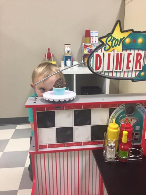 A child peeks over a toy diner counter with a cupcake on display, surrounded by colorful condiment bottles.