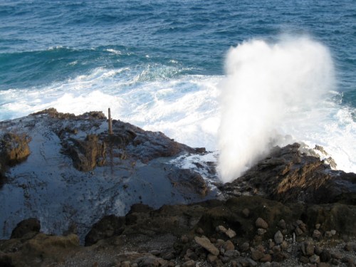 Waves crashing against rocky shore, creating a spray of water against a backdrop of the ocean.