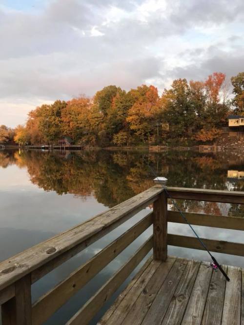 A serene lake view at sunset, with colorful autumn trees reflecting on the water from a wooden dock.
