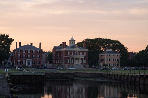 Historic buildings by a calm waterfront at sunset, reflecting warm colors in the water.