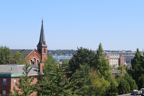 A scenic view of a town featuring a church steeple, trees, and a glimpse of the waterfront in the background.
