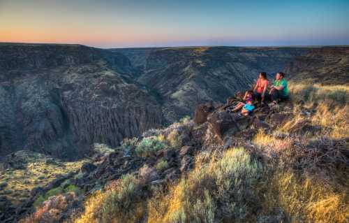 A group of people sitting on rocks overlooking a vast canyon at sunset, surrounded by rugged terrain and vegetation.