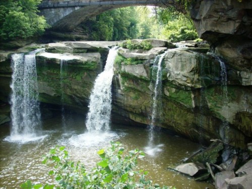 A serene scene of waterfalls cascading over rocky cliffs, surrounded by lush greenery and a bridge overhead.