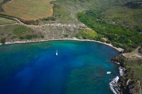 Aerial view of a serene bay with a sailboat, surrounded by lush greenery and rocky coastline.