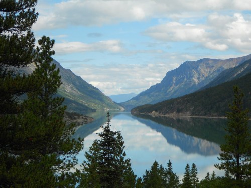 A serene lake surrounded by mountains, reflecting the sky and trees, under a partly cloudy blue sky.