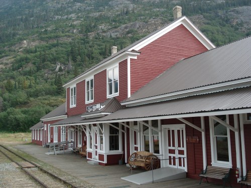 Historic red train station with a sloped roof, surrounded by lush green mountains and a gravel path.