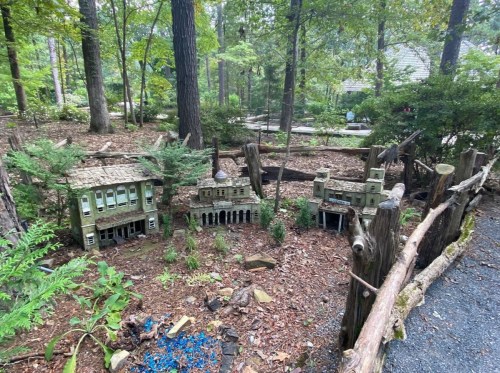 Miniature buildings nestled among trees in a wooded area, surrounded by natural foliage and a rustic wooden fence.