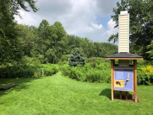 A nature trail with a sign about local wildlife, surrounded by lush greenery and trees under a partly cloudy sky.