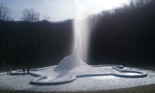 A large, snowy fountain erupts in a forested area, with people standing nearby and mountains in the background.