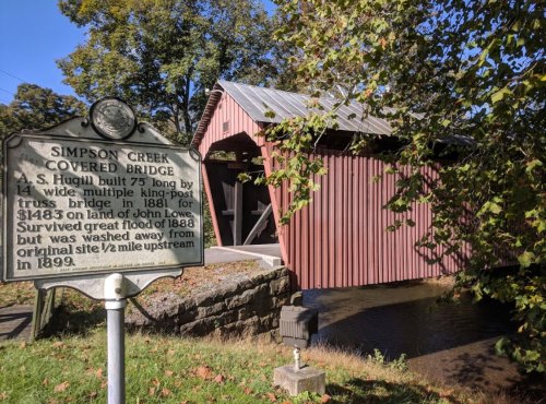A red covered bridge with a historical marker nearby, surrounded by trees and reflecting in the creek below.
