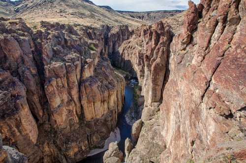 Aerial view of a deep canyon with steep rock walls and a winding river at the bottom, surrounded by rugged terrain.