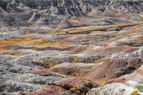 Vast, colorful badlands with layered hills and valleys in shades of gray, purple, and yellow under a clear sky.