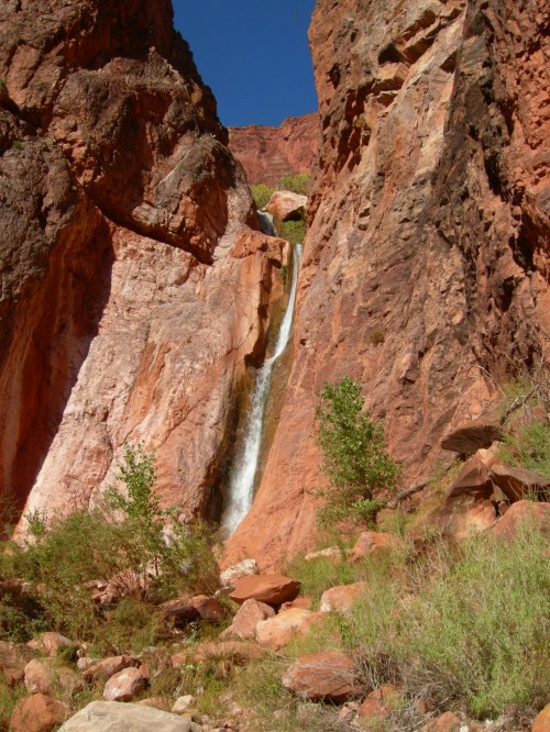 A waterfall cascading down red rock formations, surrounded by greenery under a clear blue sky.