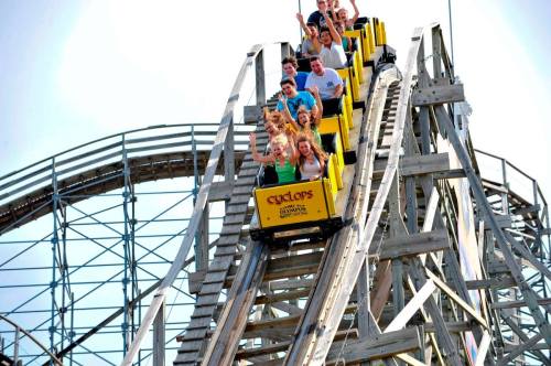 A group of excited riders on a yellow roller coaster, descending a steep track with hands raised in the air.