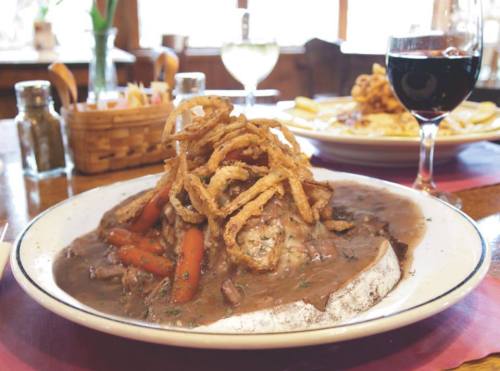 A plate of beef with gravy, topped with crispy onions and carrots, served with a glass of red wine in a cozy setting.