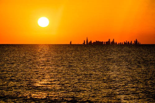 Sunset over a calm ocean, with a city skyline silhouetted against the vibrant orange sky.