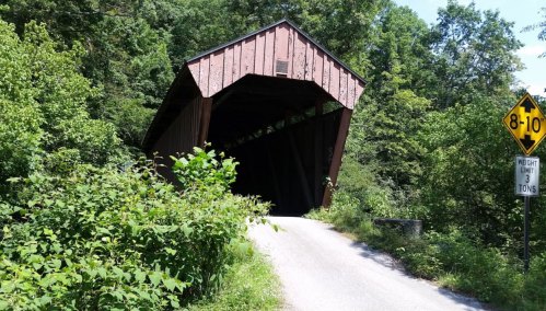 A tilted covered bridge surrounded by lush greenery, with a weight limit sign nearby.