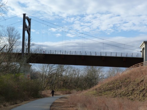 A suspension bridge spans above a walking path, surrounded by trees and a cloudy sky.