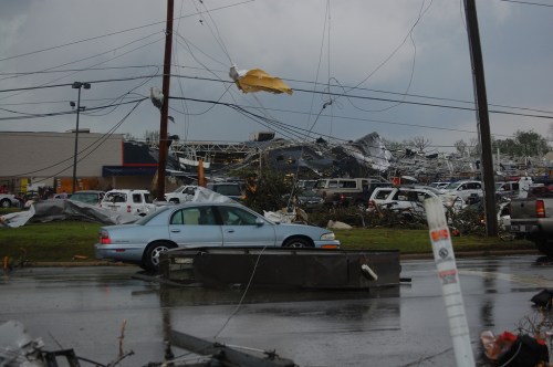A damaged area after a storm, featuring downed power lines, debris, and a car amidst destruction.