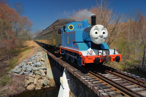 A cheerful blue train, resembling Thomas the Tank Engine, crosses a bridge with a vintage passenger car behind it.