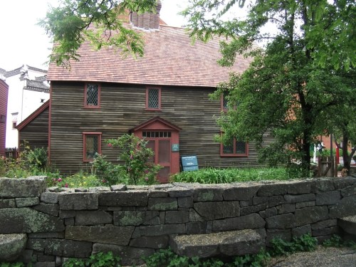 A rustic wooden house with a red roof, surrounded by greenery and a stone wall, set in a quaint outdoor area.