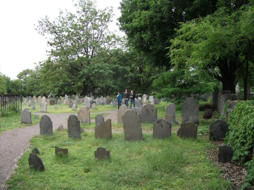 A cemetery with numerous gravestones surrounded by trees and grass, with two people walking along a path.