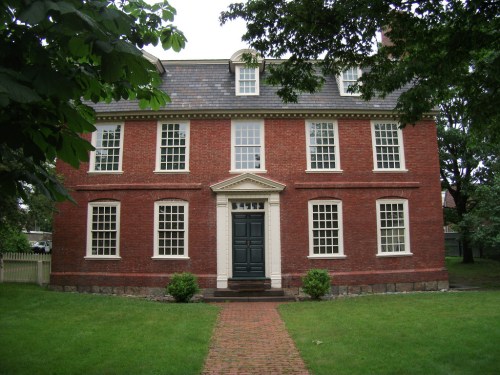 A large red brick house with white trim, featuring multiple windows and a central front door, surrounded by green grass.