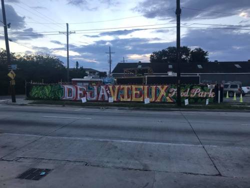 Colorful mural reading "Deja Vieux" on a fence, with a cloudy sky and street in the foreground.