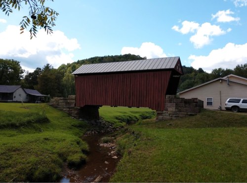 A red covered bridge spans a small stream, surrounded by green grass and trees under a blue sky with clouds.
