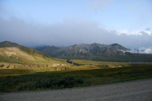 A scenic view of mountains under a cloudy sky, with green hills and a gravel road in the foreground.