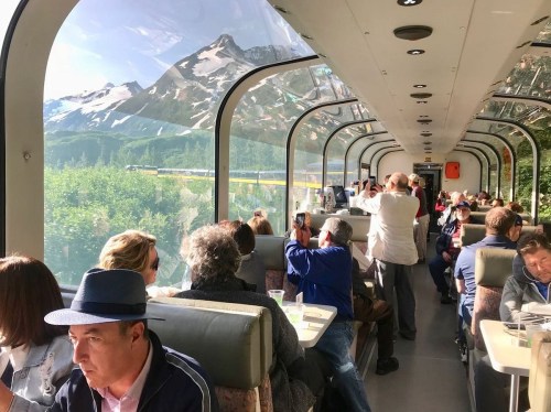 Passengers enjoy scenic views from a glass-roofed train, surrounded by mountains and greenery.