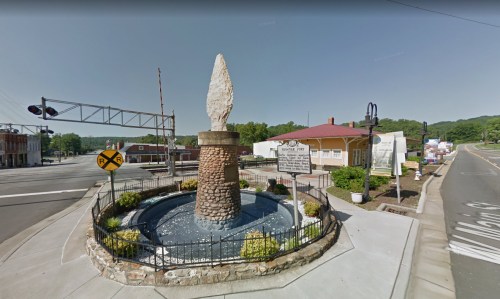 A stone monument with a large pointed sculpture, surrounded by a small fountain and greenery, near a railroad crossing.