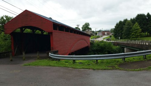 A red covered bridge spans a small creek, with a road and trees in the background under a cloudy sky.