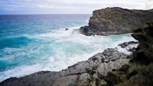 Waves crash against rocky cliffs under a cloudy sky, with a vibrant blue ocean in the background.