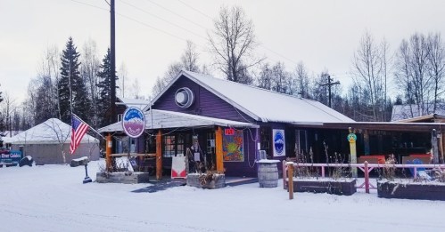 A cozy purple building with an American flag, surrounded by snow and trees, featuring colorful signs and an open sign.