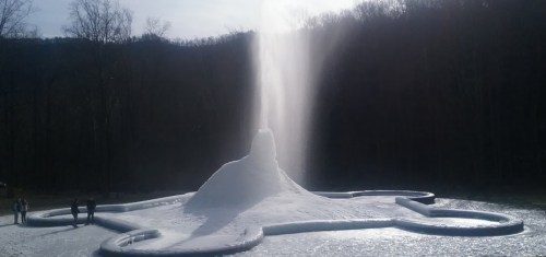 A large, snowy fountain erupts in a park, surrounded by trees and a walking path with people nearby.
