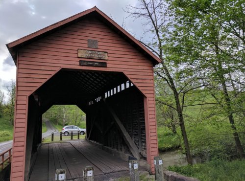 A red covered bridge with a plaque, surrounded by greenery and a road, with a car parked nearby.