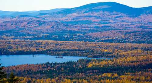 A scenic view of a colorful forested landscape with a lake, surrounded by mountains under a clear sky.