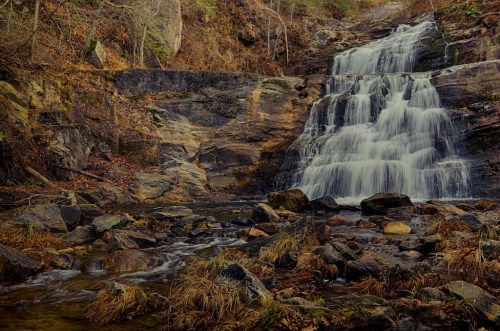 A serene waterfall cascades over rocky cliffs, surrounded by autumn foliage and a rocky riverbed.