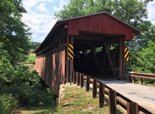 A red covered bridge surrounded by greenery, with a gravel road leading to it and warning signs on either side.